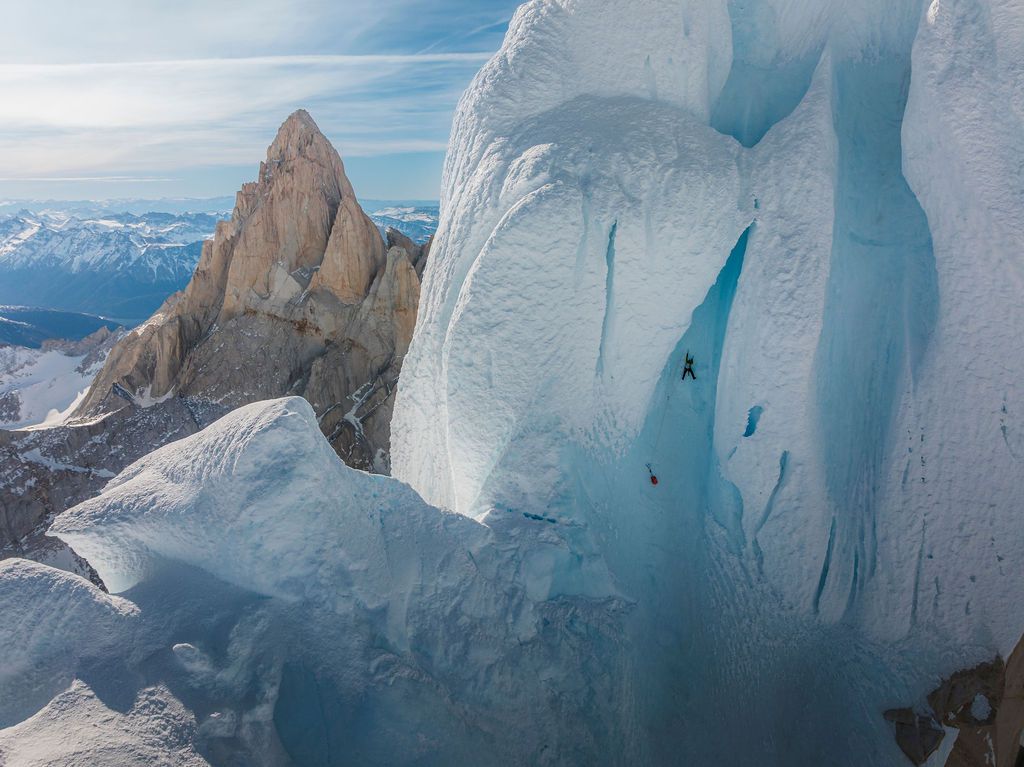 Two climbers on a massive blue ice wall with a sharp granite spire rising behind them in Patagonia.