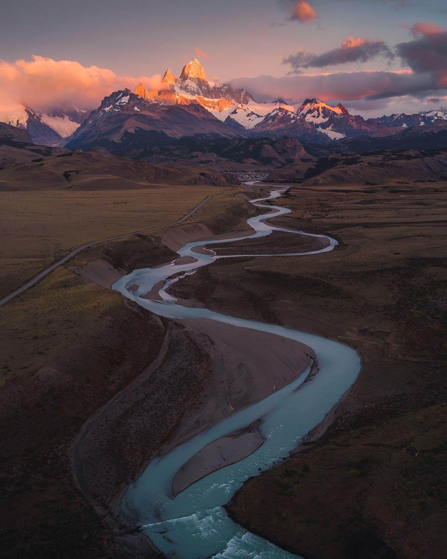 Aerial view of winding turquoise river snaking through Patagonian steppe toward glowing Fitz Roy at sunset.