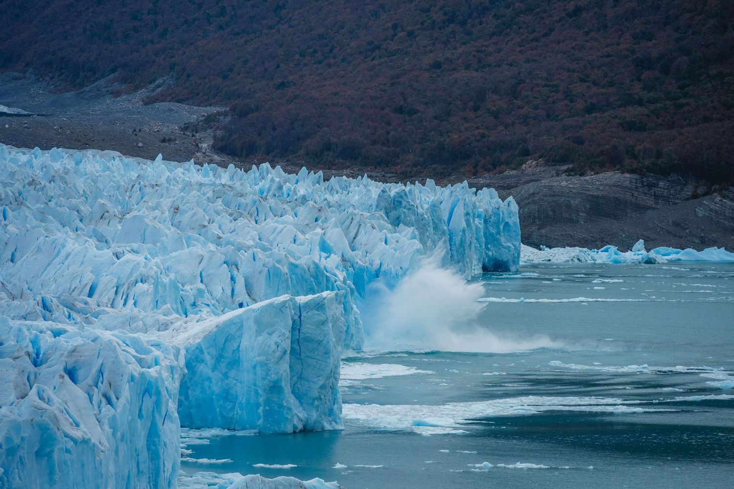 Massive blue glacier calving into turquoise waters with ice chunks splashing, surrounded by rocky mountains in Patagonia.