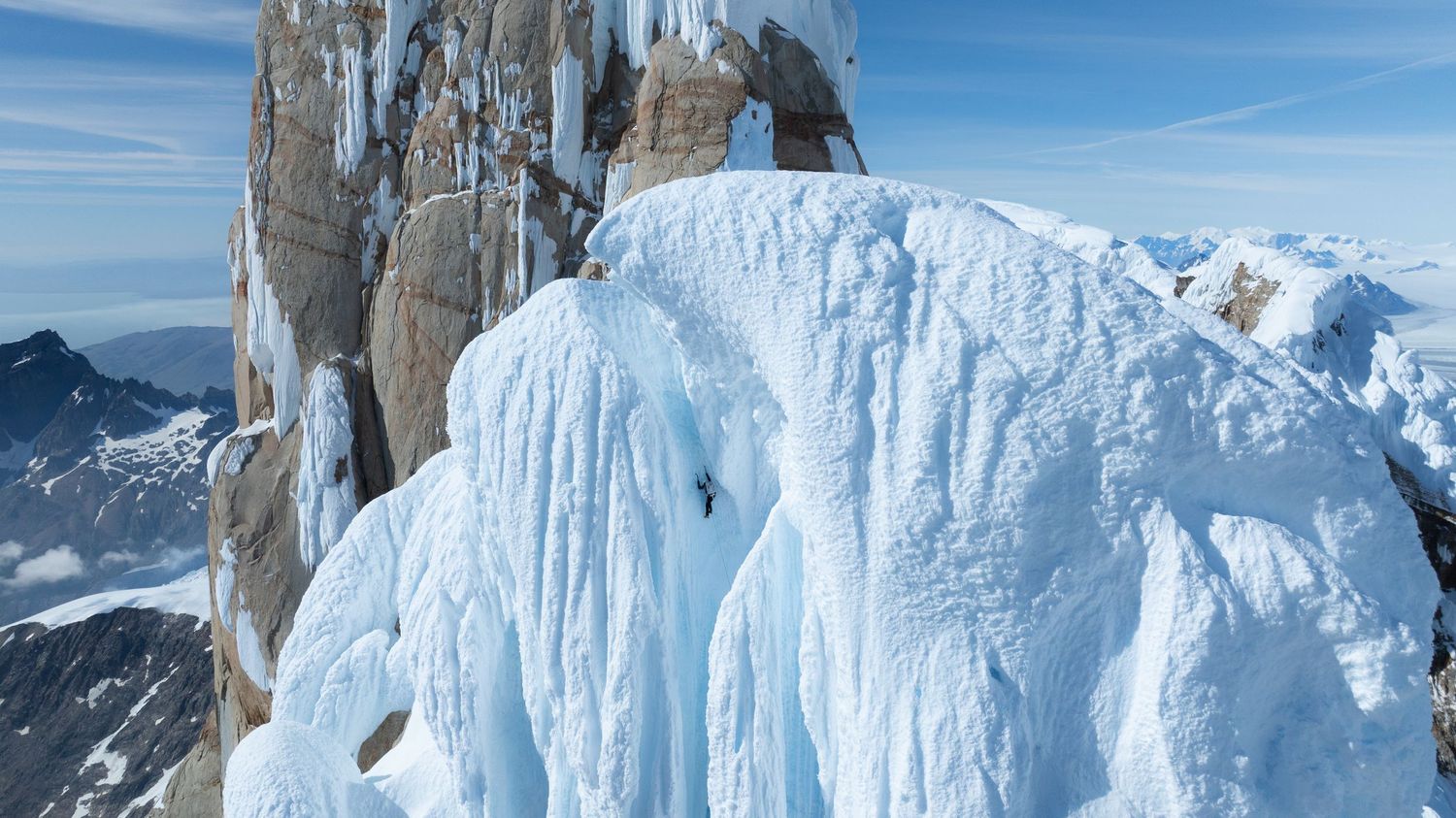 Ice climber scaling a dramatic frozen ice formation on a rocky mountain peak under a clear blue sky.