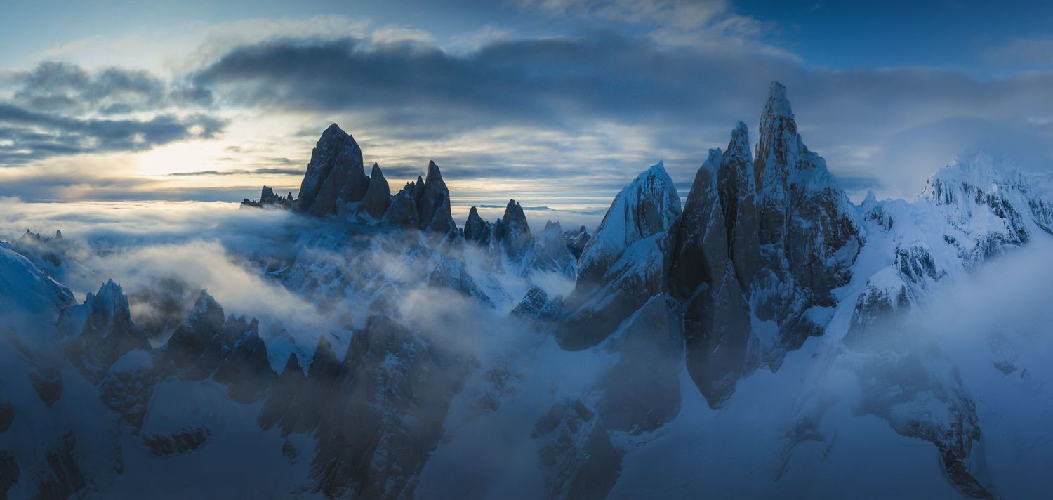 Snow-capped jagged mountain peaks emerge through dramatic clouds at twilight in a stunning Patagonian landscape panorama.