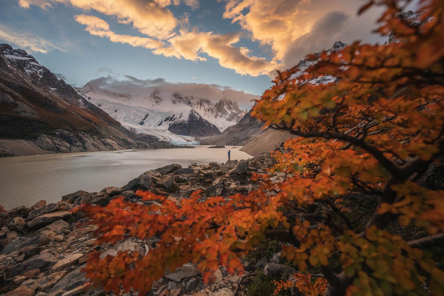 Vibrant autumn foliage frames a lone figure by a glacial lake below Cerro Torre, Patagonia.