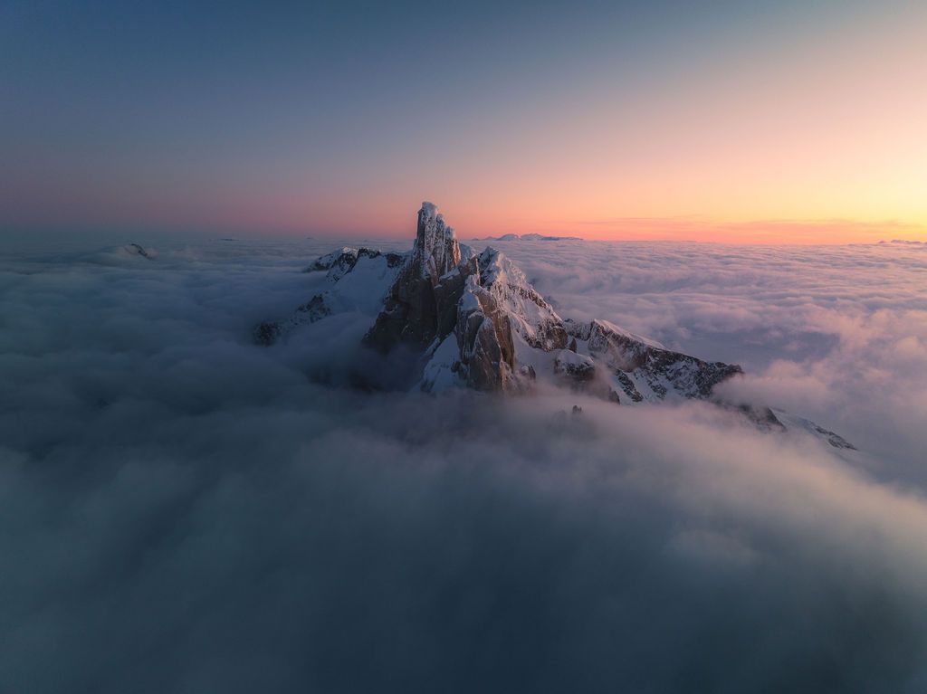 A solitary snow-dusted mountain peak emerges above a vast sea of clouds during a colorful twilight sunset.