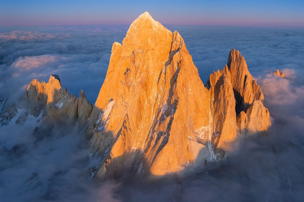 Close-up aerial view of Cerro Torre peak glowing golden at sunrise above a sea of clouds in Patagonia.