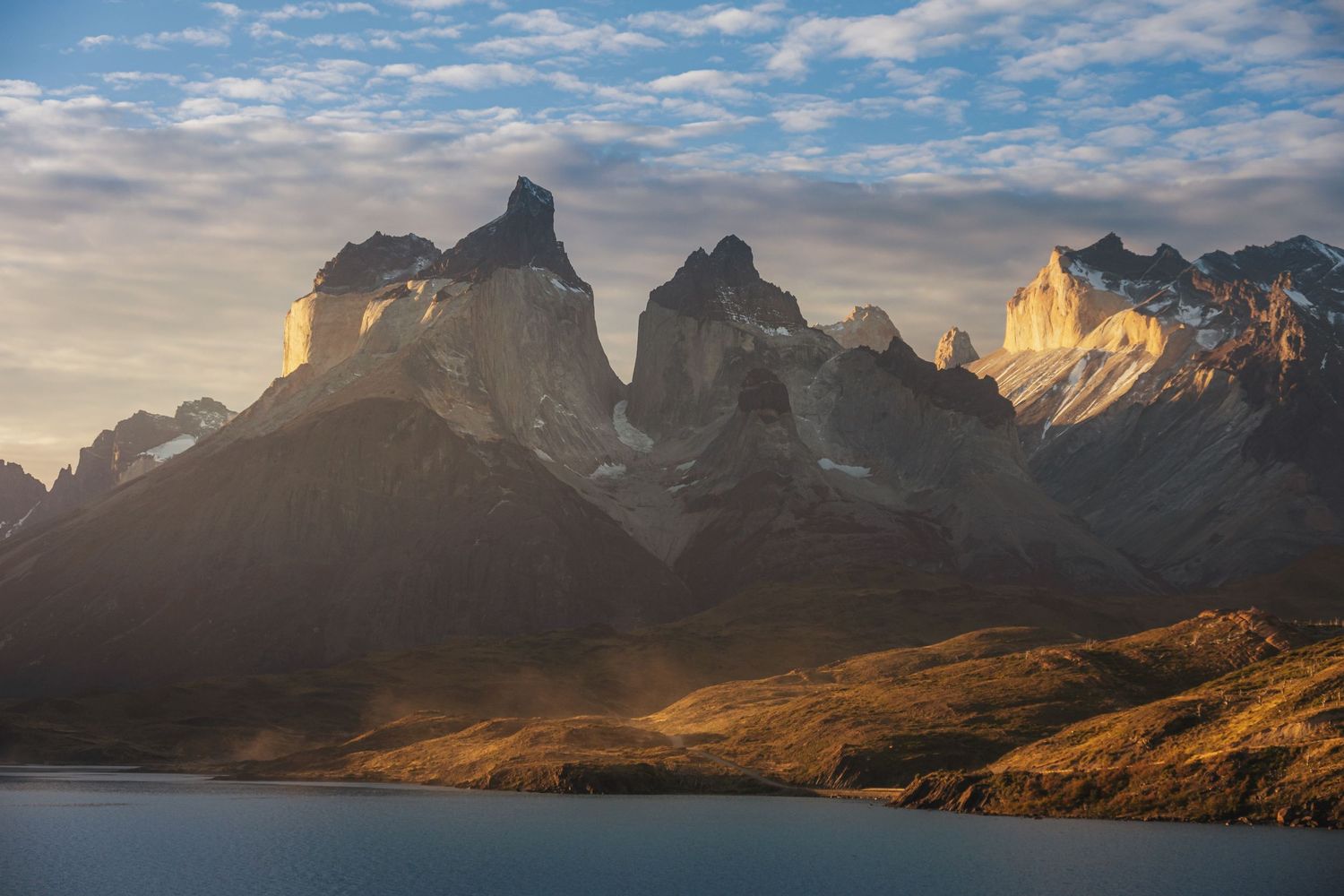 Golden sunrise illuminates jagged mountain peaks above a serene lake in Torres del Paine National Park, Patagonia.
