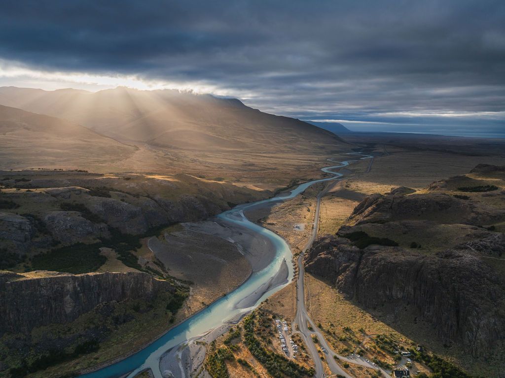 Aerial view of a turquoise river winding through a vast Patagonian valley with dramatic sunrays breaking through clouds.