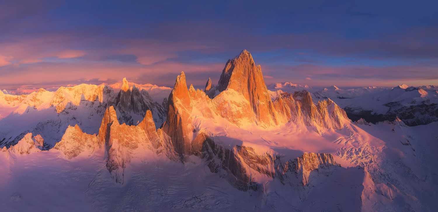 Panoramic aerial view of jagged snow-covered Fitz Roy mountain peaks glowing orange at sunset in Patagonia.