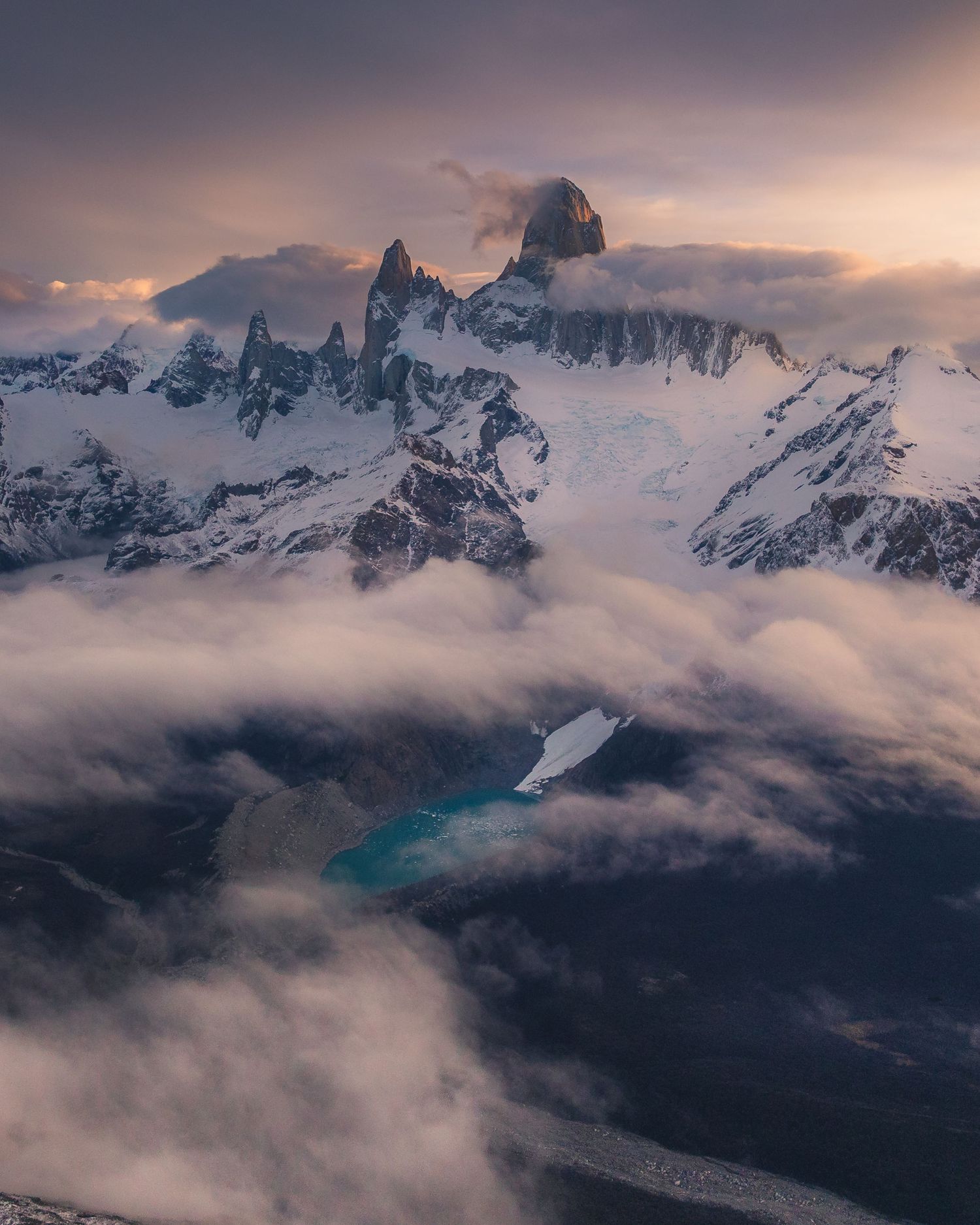 Snow-capped Fitz Roy peaks emerge through dramatic clouds at sunset in Patagonia, with a turquoise glacial lake below.