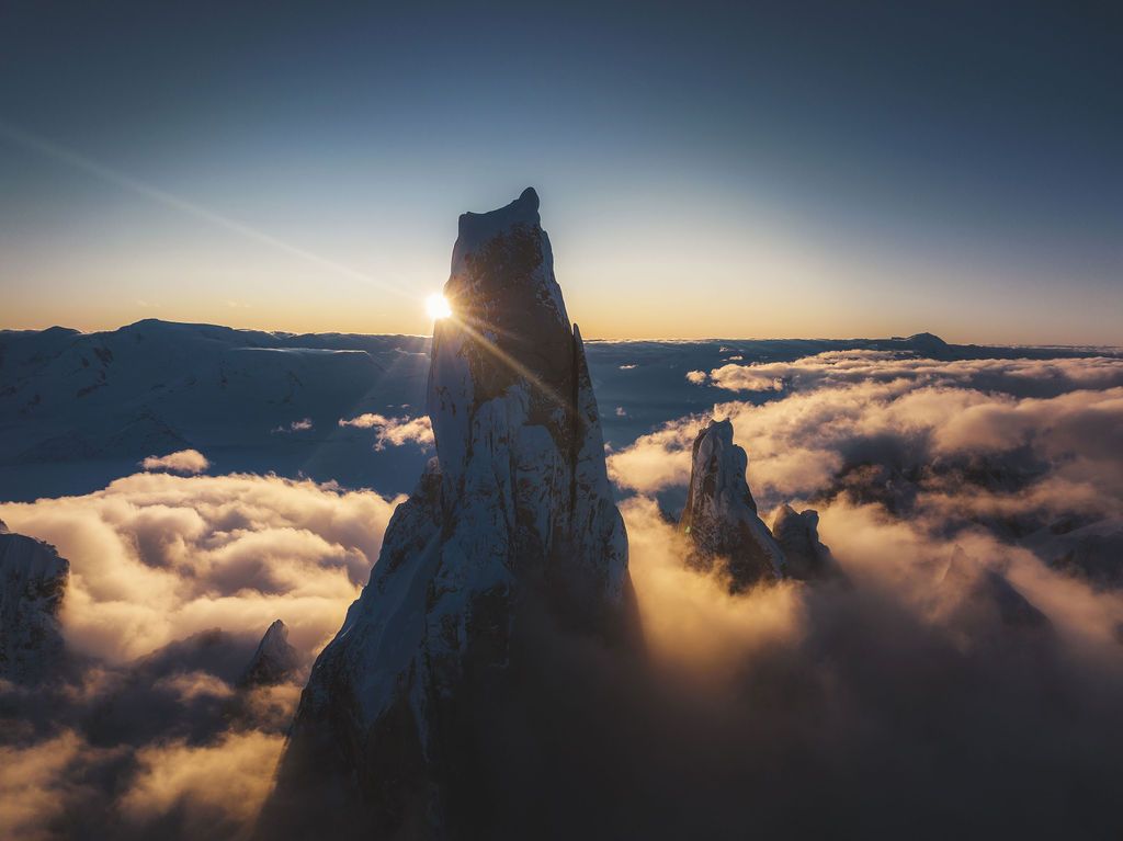 A brilliant sunstar bursts between jagged snow-covered rock spires rising above a golden cloud sea at sunset.