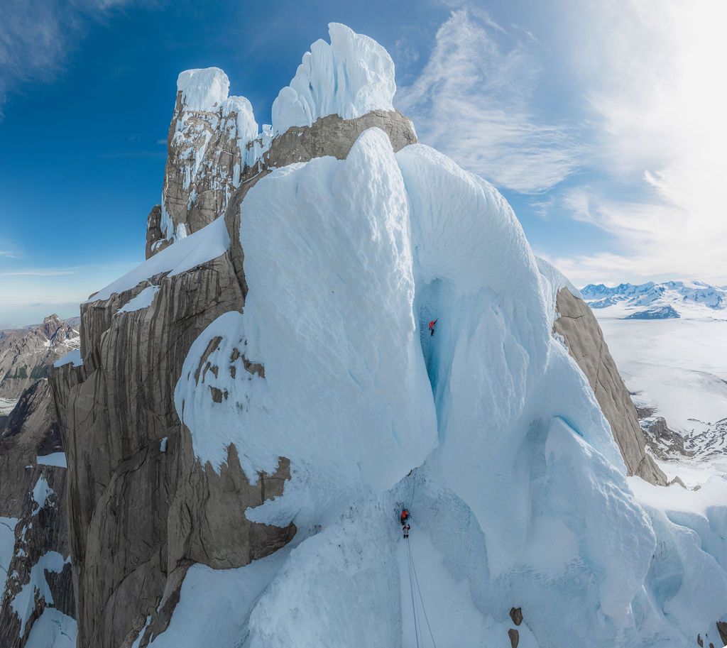 Two climbers ascending a giant ice mushroom on a rocky Patagonian peak with vast cloud-covered landscape below.