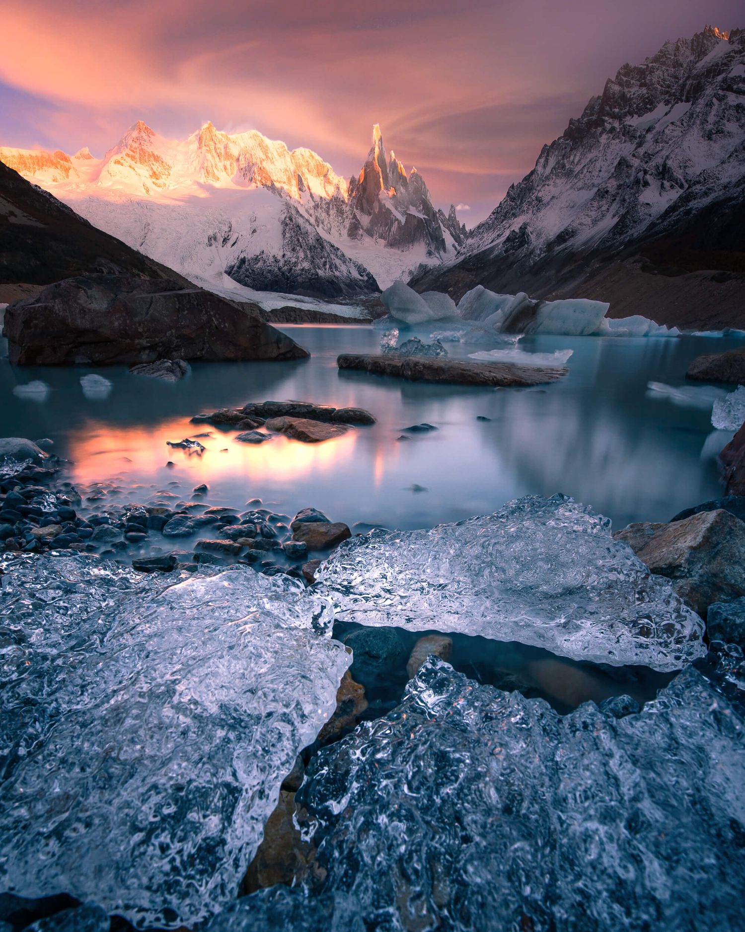 Icy glacial lake with frozen chunks reflecting pink sunset sky and snow-capped Cerro Torre in Patagonia.