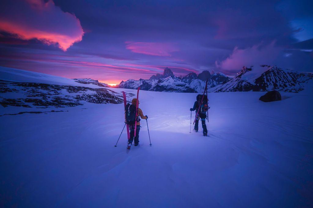 Two mountaineers ski across a snowy glacier at twilight with dramatic purple and pink sky in Patagonia.