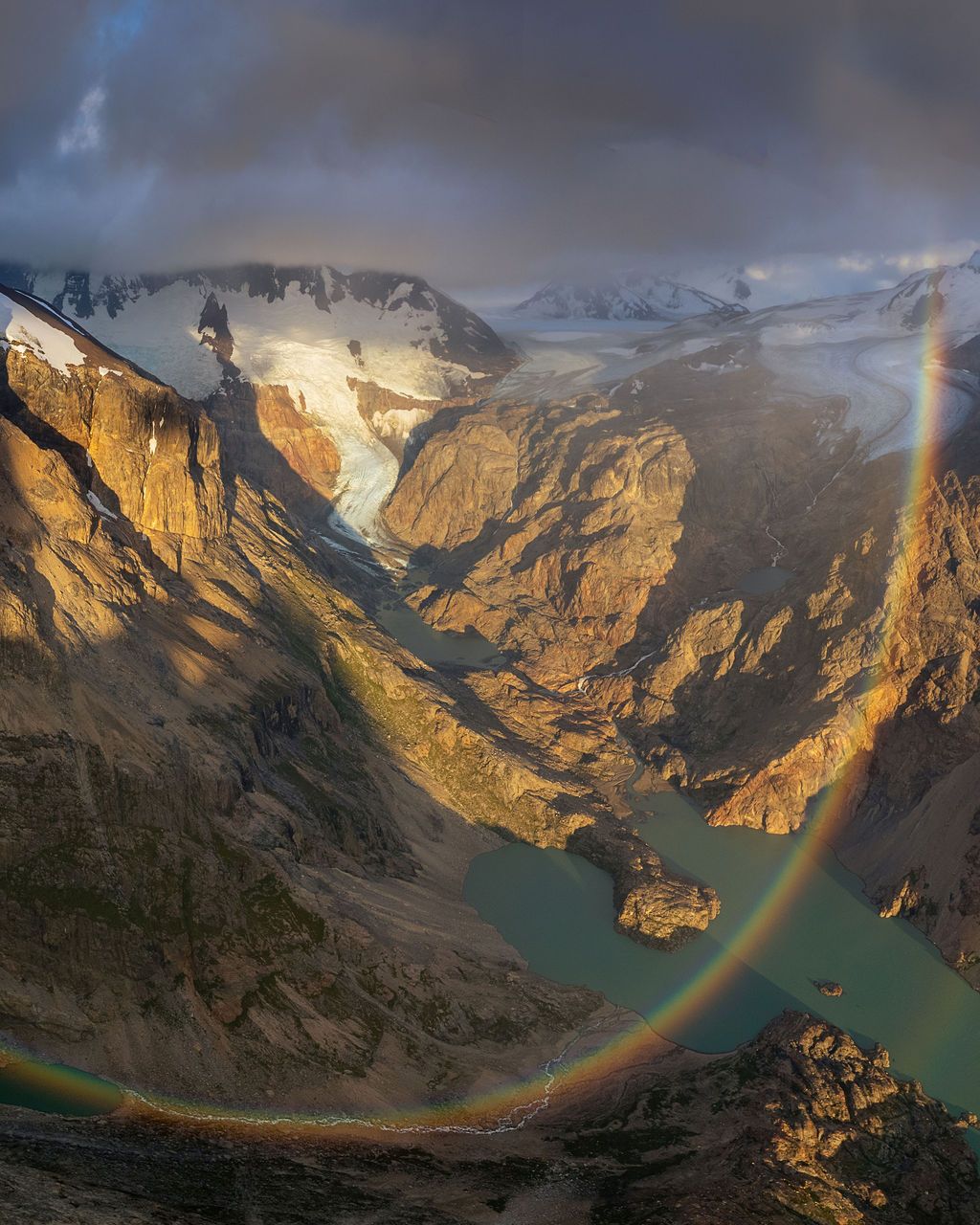 A vibrant rainbow arcs over a glacial mountain valley with turquoise lake and dramatic stormy light.