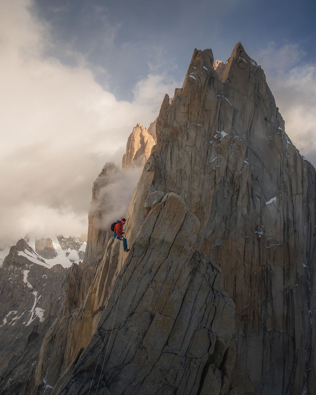 Climber scaling jagged rocky peaks in Patagonia with dramatic clouds and snow-capped mountains in background.