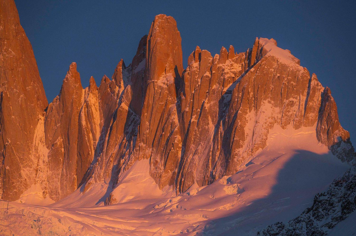 Dramatic orange alpenglow illuminates jagged granite spires of Cerro Torre range at sunrise in Patagonia.