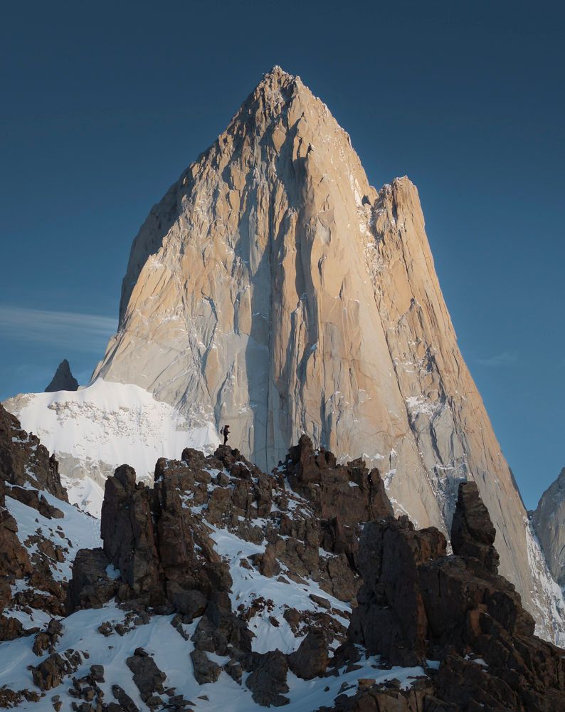 Tiny climber stands below the towering granite spire of Mount Fitz Roy at sunrise.