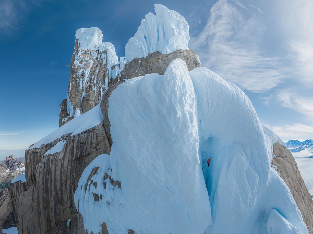 Lone climber scaling a towering ice and rock formation on a Patagonian mountain summit under blue skies.