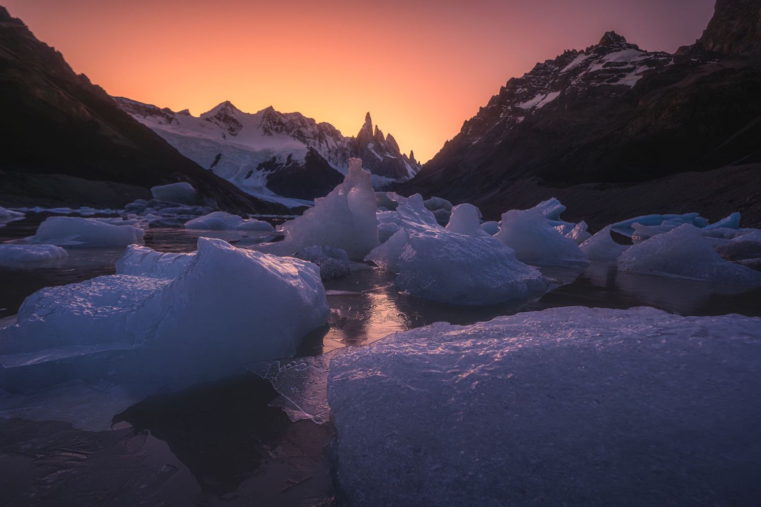 Dramatic sunset over icy Patagonian landscape with jagged snow-capped peaks glowing orange and blue ice chunks in foreground.