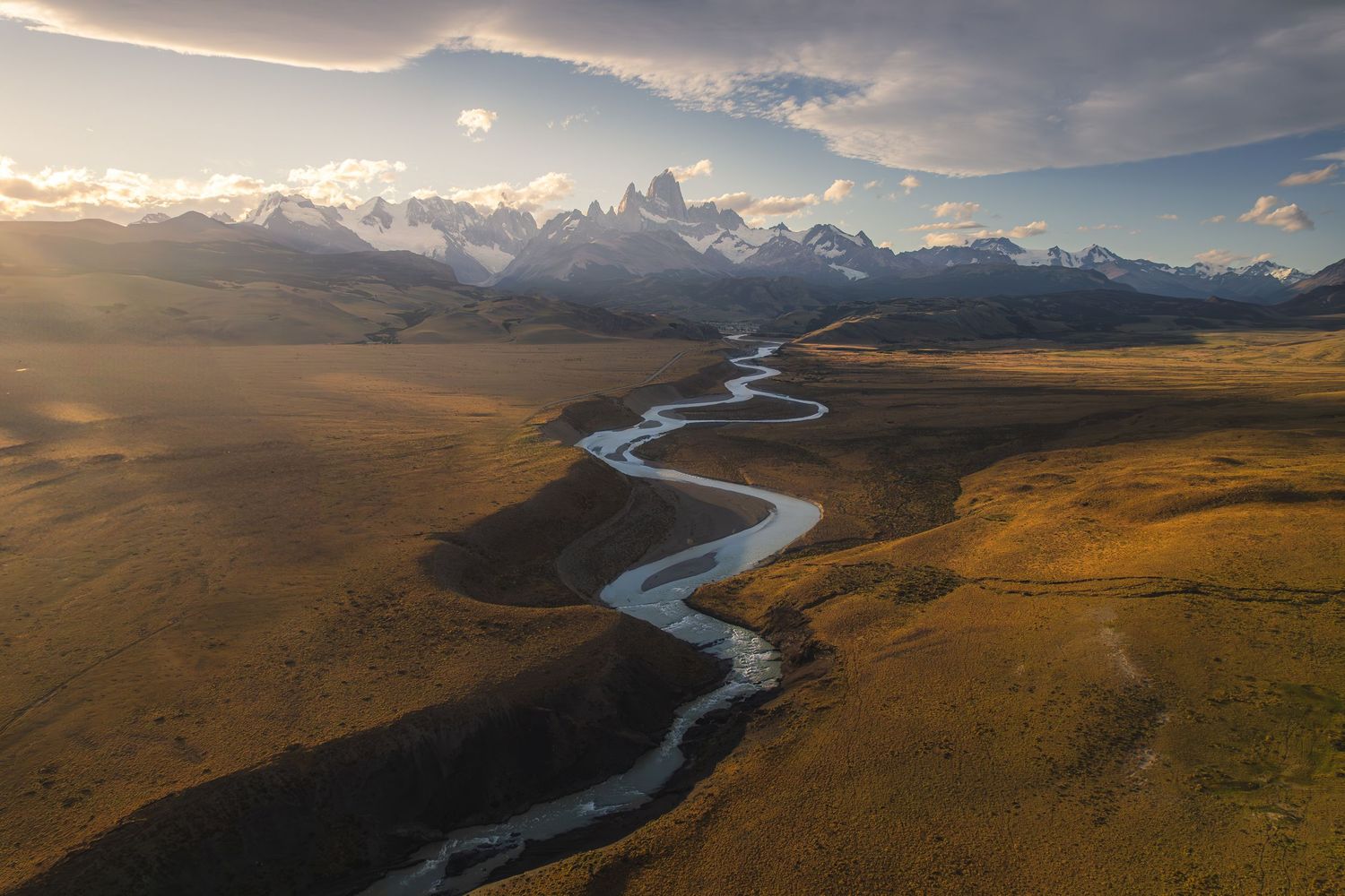 Aerial view of a winding river through golden Patagonian plains with snow-capped Andes mountains at sunset.