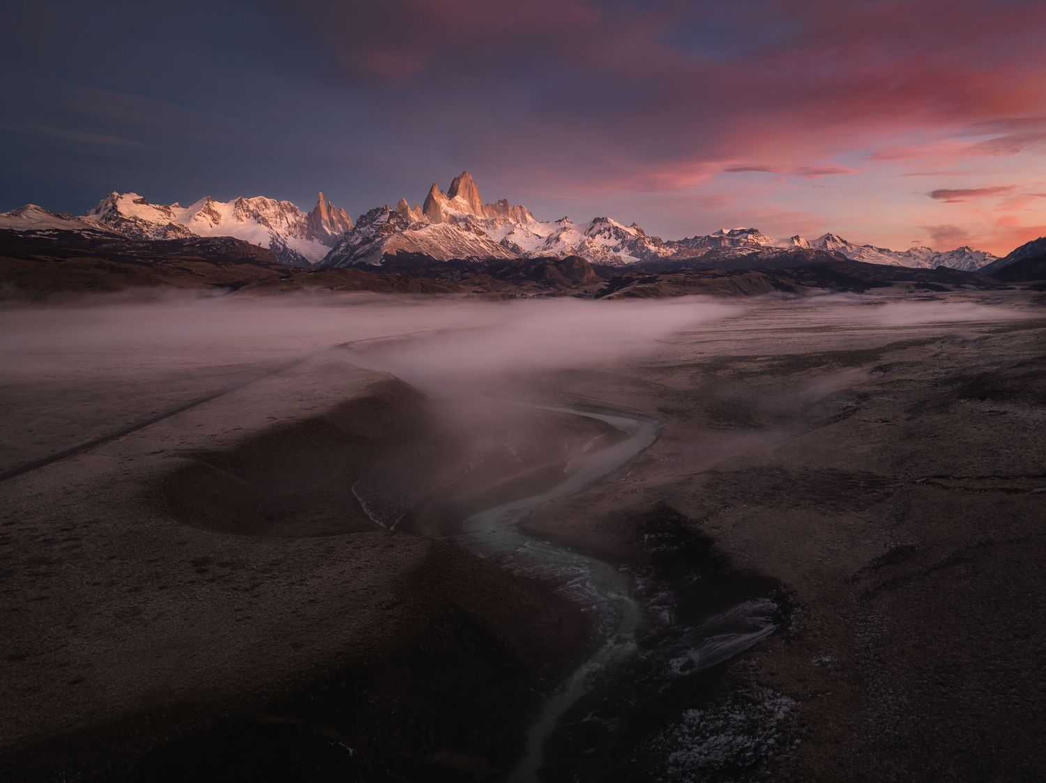 Aerial view of Fitz Roy mountain at sunset with misty valley, winding river, and dramatic pink sky in Patagonia.