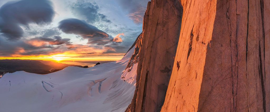 Climbers scale a towering red granite wall at sunset with vast snowy plateau glowing in the background.