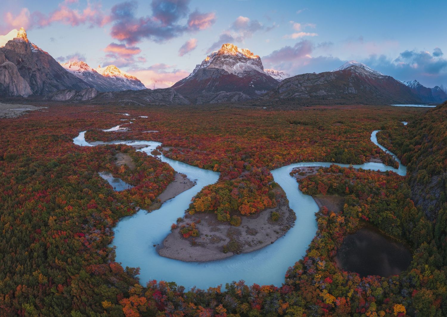 Aerial view of a winding turquoise river through autumn forest with snow-capped Patagonian peaks at sunset.