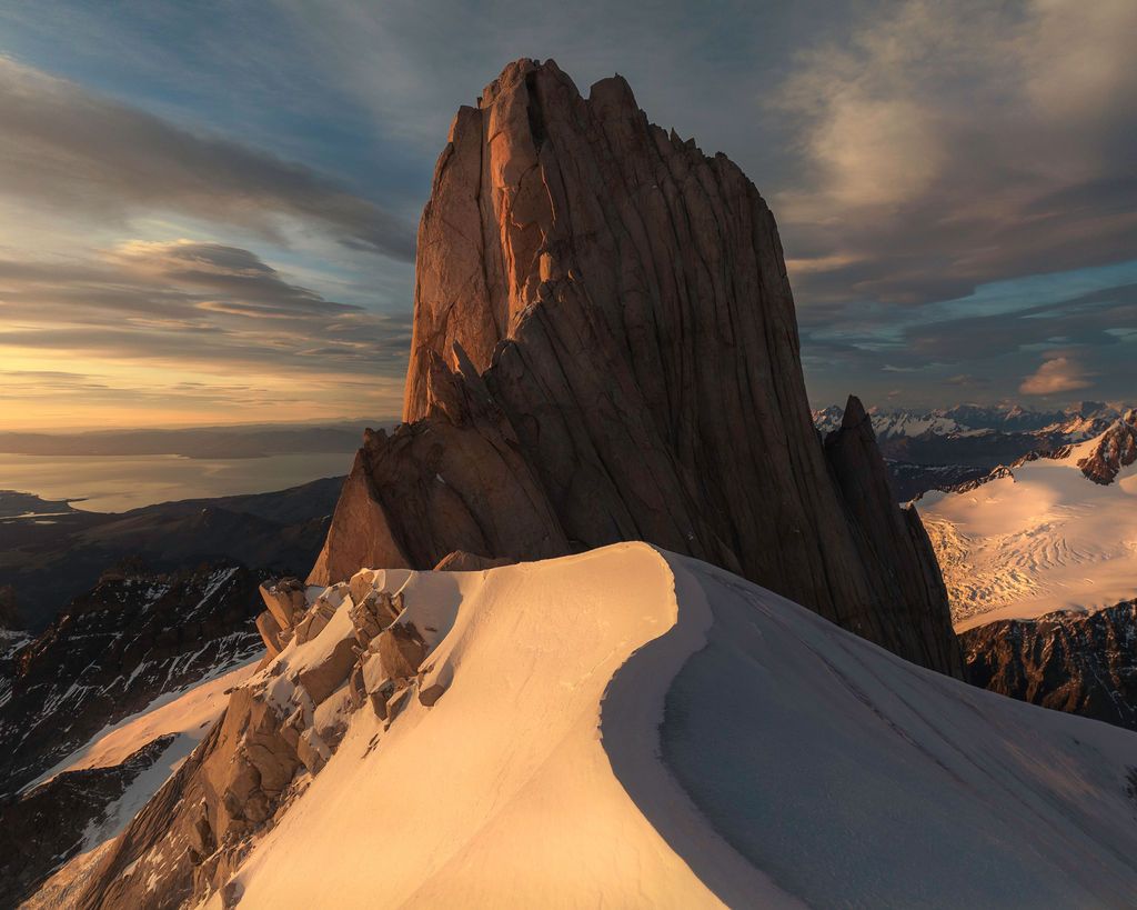 Golden sunrise light illuminates a massive granite monolith rising above a snow ridge in Patagonia.