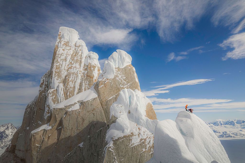 Mountaineer standing on an ice-covered rocky summit with vast snowy peaks stretching to the horizon.