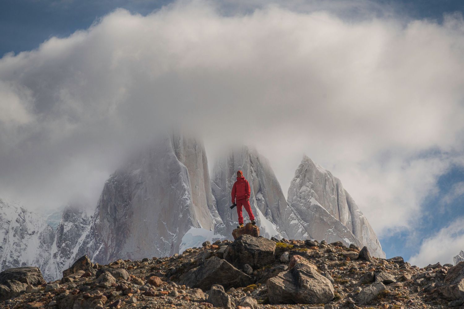Hiker in red jacket stands on rocky terrain before snow-capped Torres del Paine peaks shrouded in clouds.