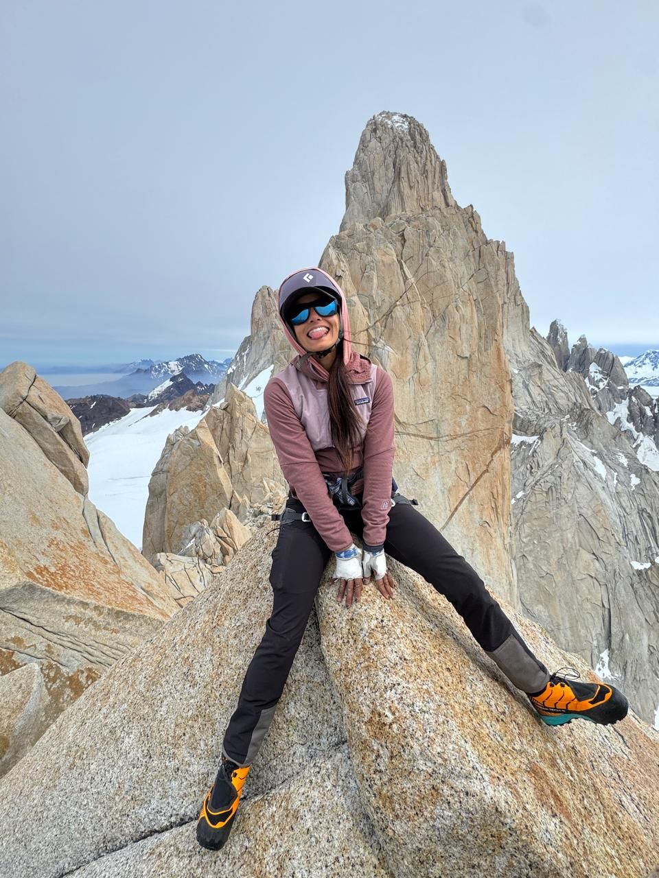 A smiling woman in climbing gear sits on a rocky summit with a dramatic jagged mountain peak in the background.