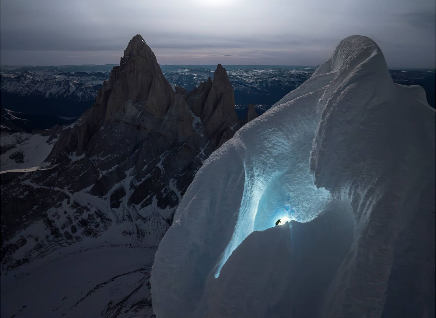 Climber explores a glowing ice cave near jagged mountain peaks at dusk in Patagonia.