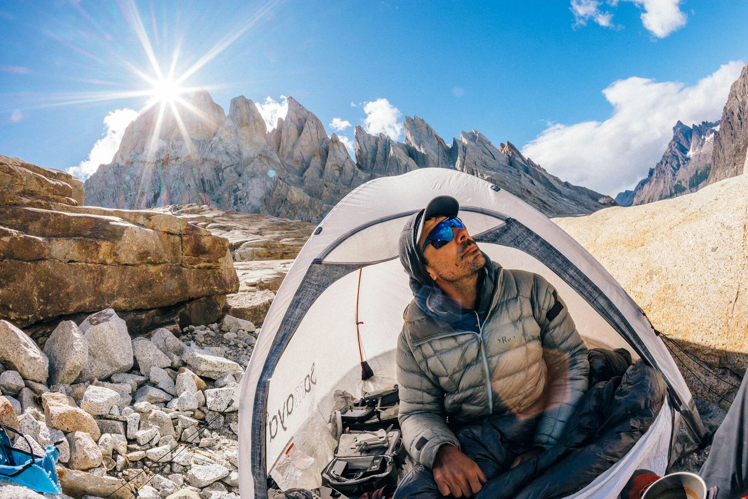 Mountain climber relaxes in tent at high altitude rocky campsite with rugged peaks under blue sky.