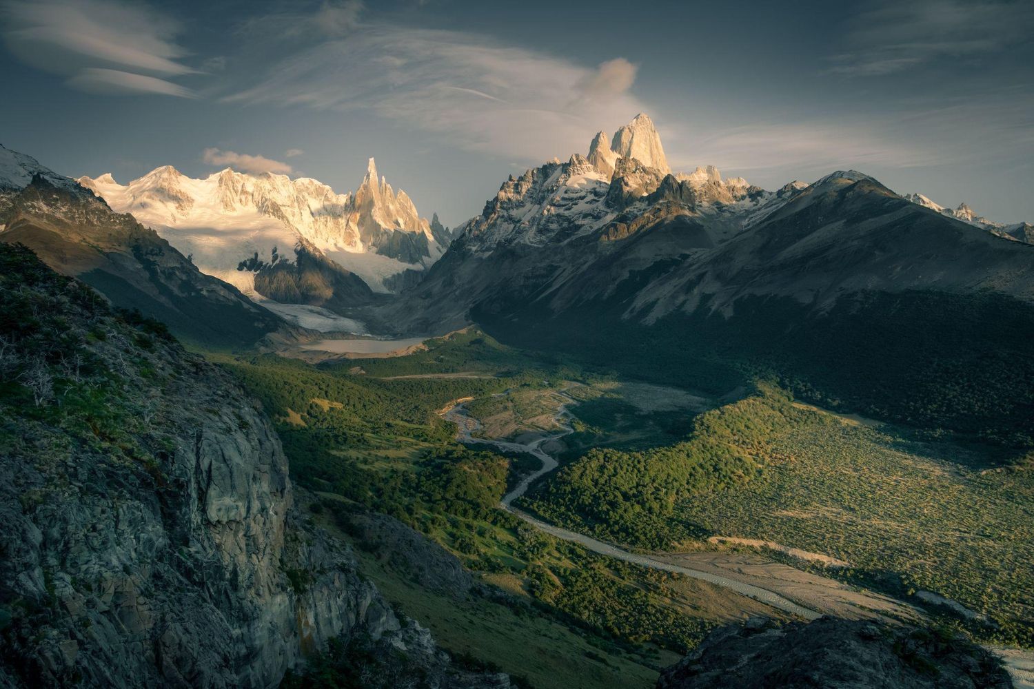 Panoramic view of Fitz Roy and Cerro Torre mountains rising above a lush green valley in Patagonia, Argentina.