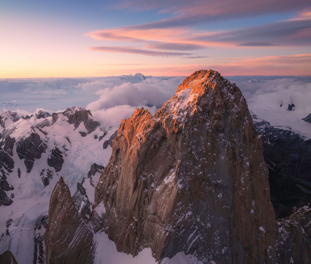 Jagged snow-dusted mountain peaks bathed in warm alpenglow rise above a dramatic pink and purple sunset sky.