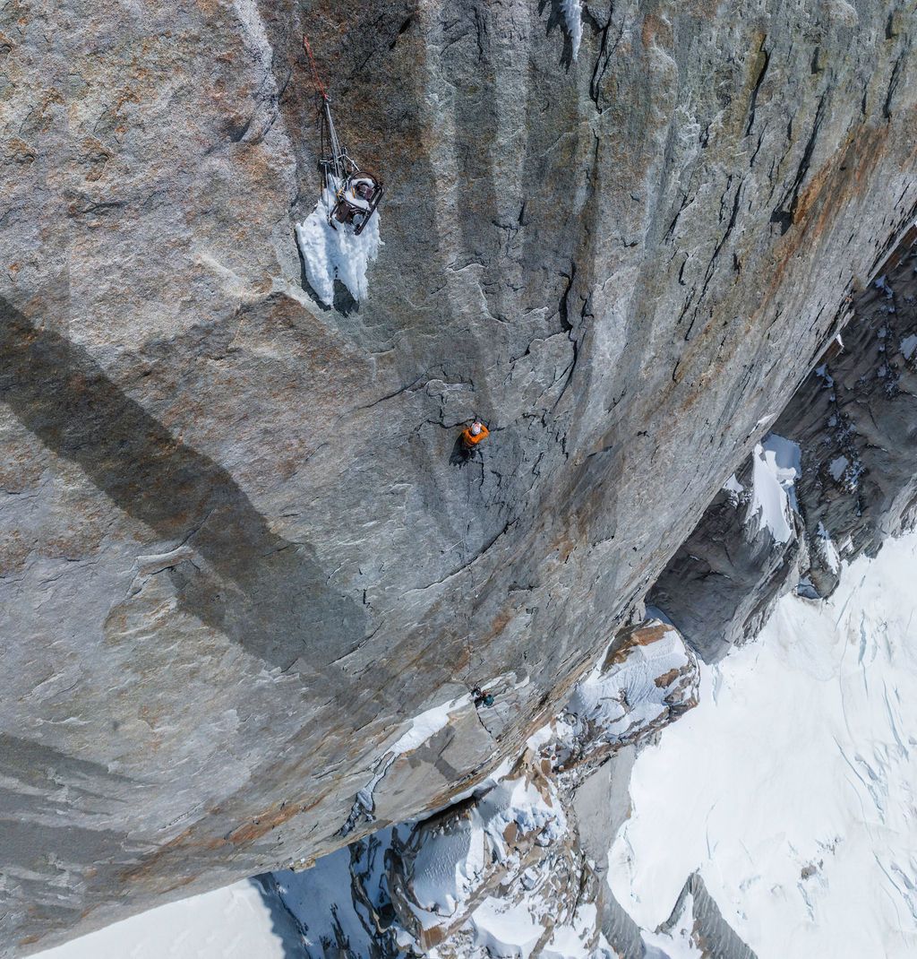 Rock climber in orange jacket scales a vertical granite wall with snowy glacier visible far below.
