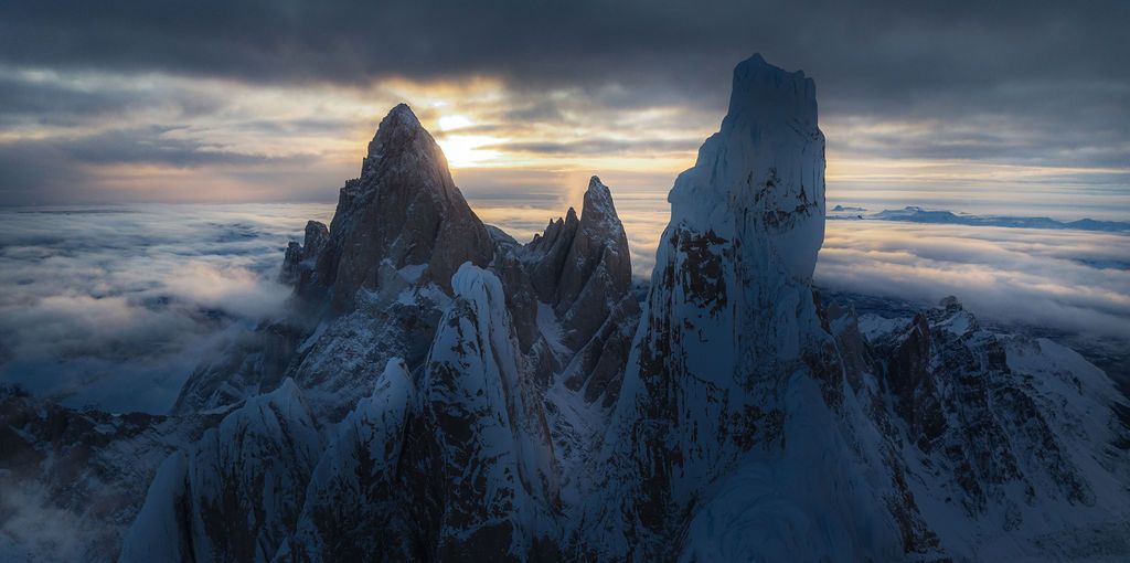 Dramatic aerial view of jagged snow-covered mountain peaks piercing through clouds at sunset in the Dolomites.