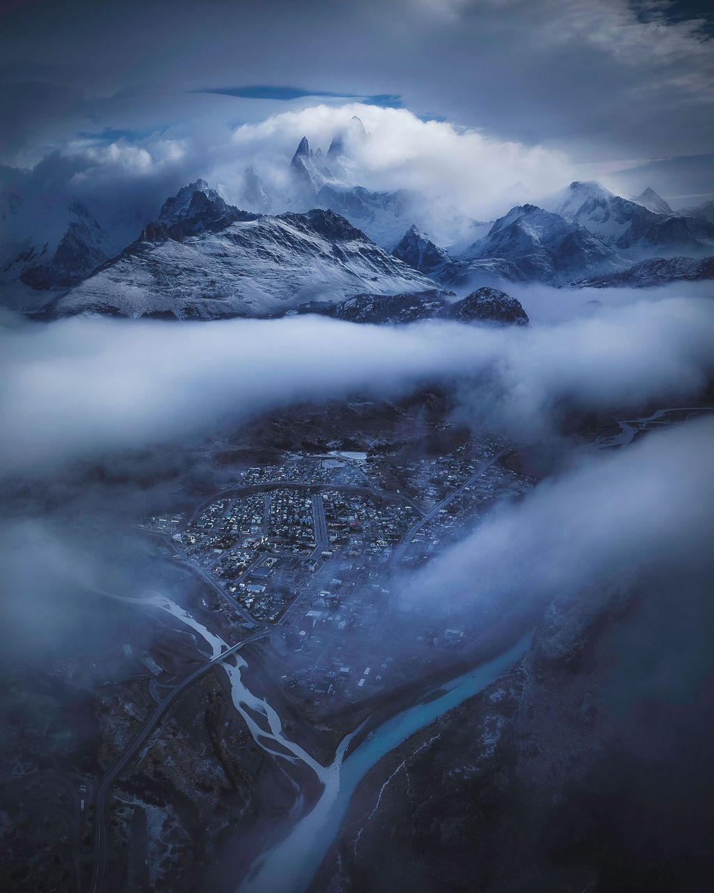 Aerial view of a snow-capped mountain town enveloped in dramatic clouds and fog, with a turquoise river winding below.