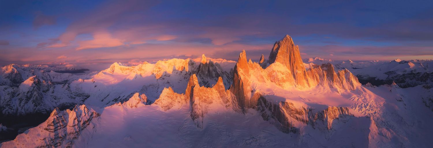 Panoramic aerial view of Fitz Roy mountain range at golden sunset, with dramatic pink and purple clouds over snow-capped Patagonian peaks.