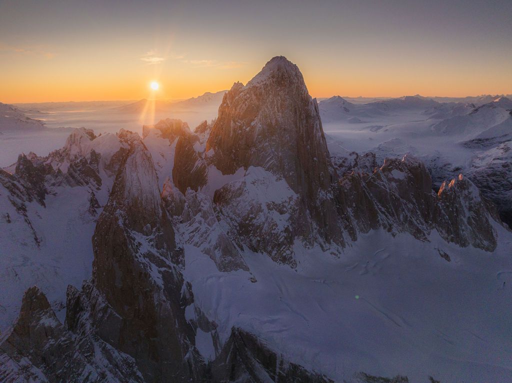 Golden sunrise bursts behind Cerro Torre's rocky spires casting warm light over snow-covered Patagonian peaks.