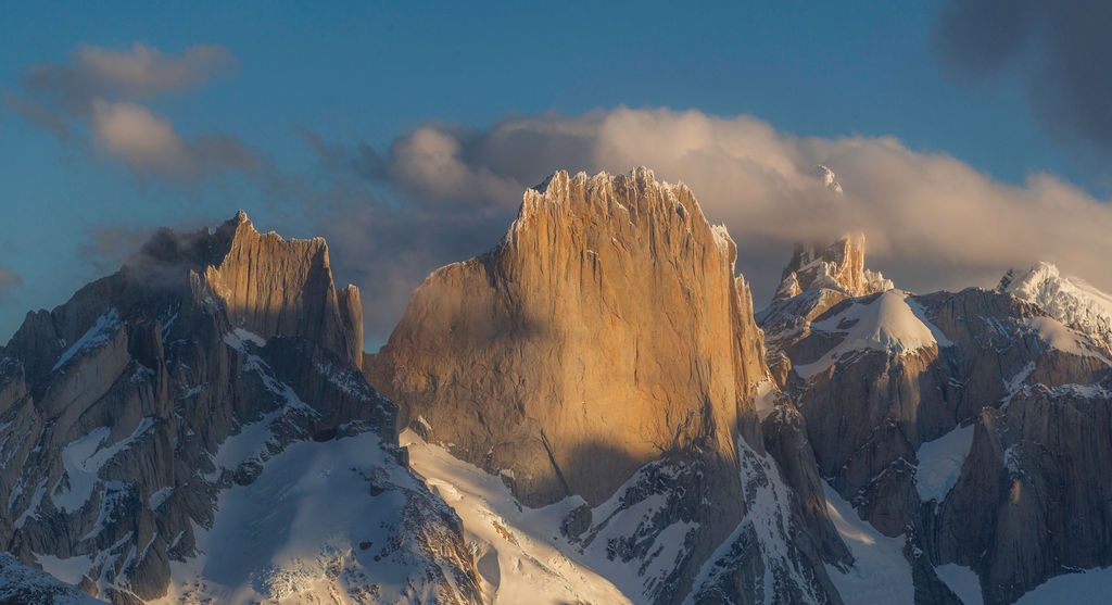 Golden sunlight illuminates the jagged snow-covered granite towers of Fitz Roy in Patagonia at sunset.