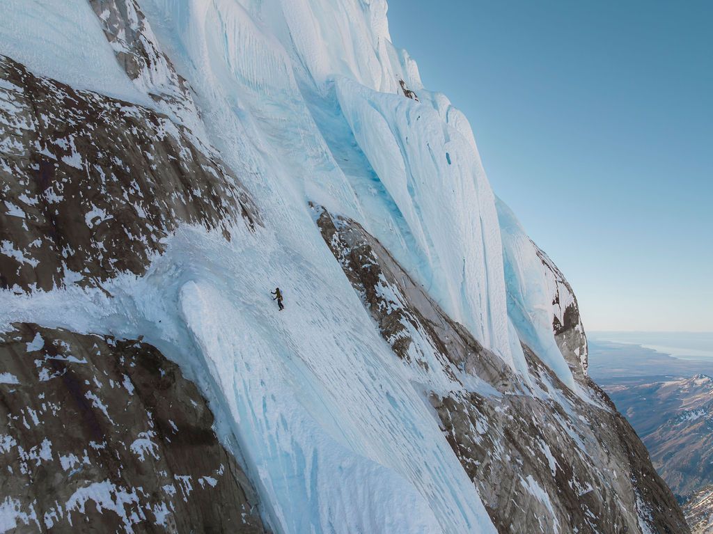 Solo climber ascending steep ice and rock face with vast snowy mountain terrain visible below.