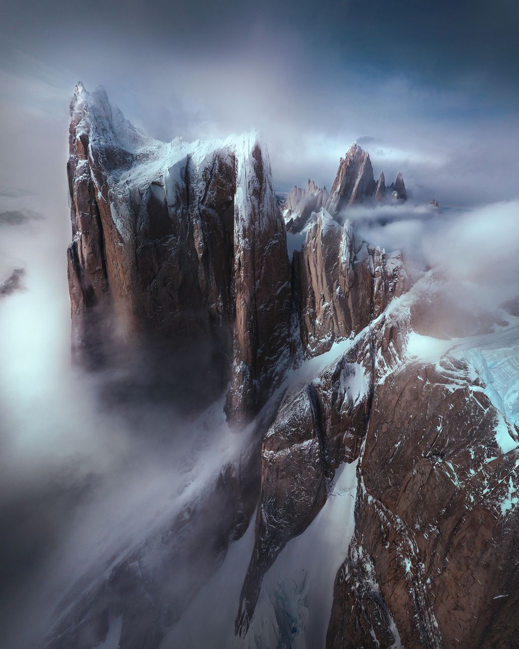 Towering jagged rocky mountain spires shrouded in moody mist and clouds in a dramatic aerial view.