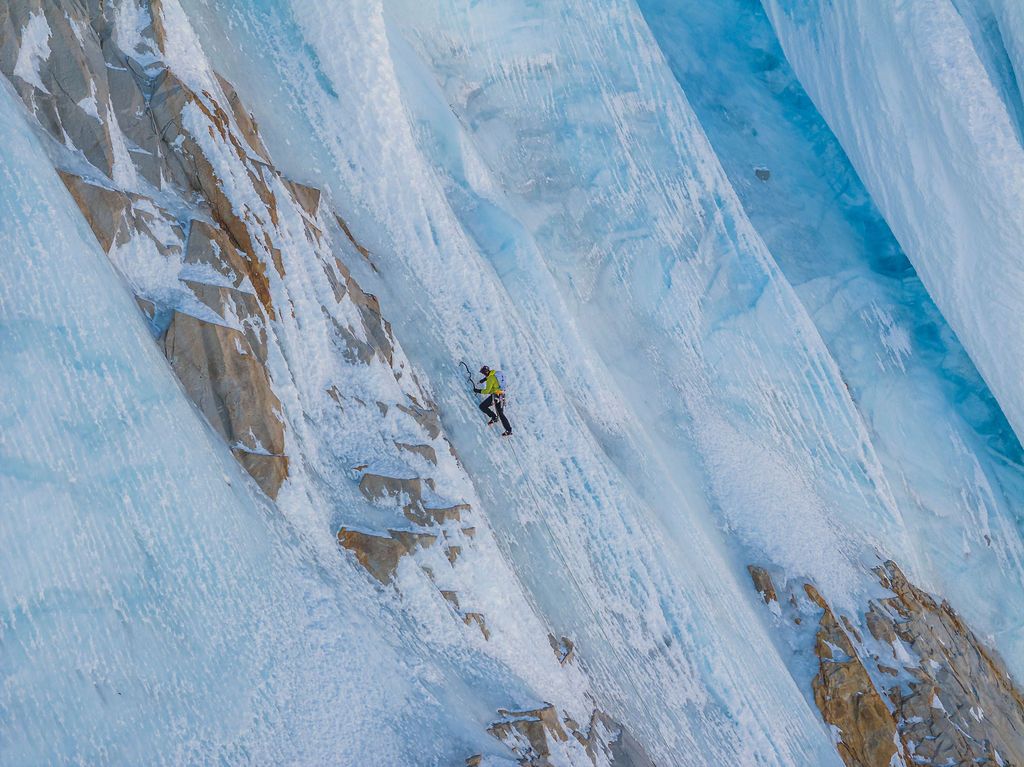 Mountaineer in yellow jacket climbing steep glacial ice wall with rocky terrain visible below.