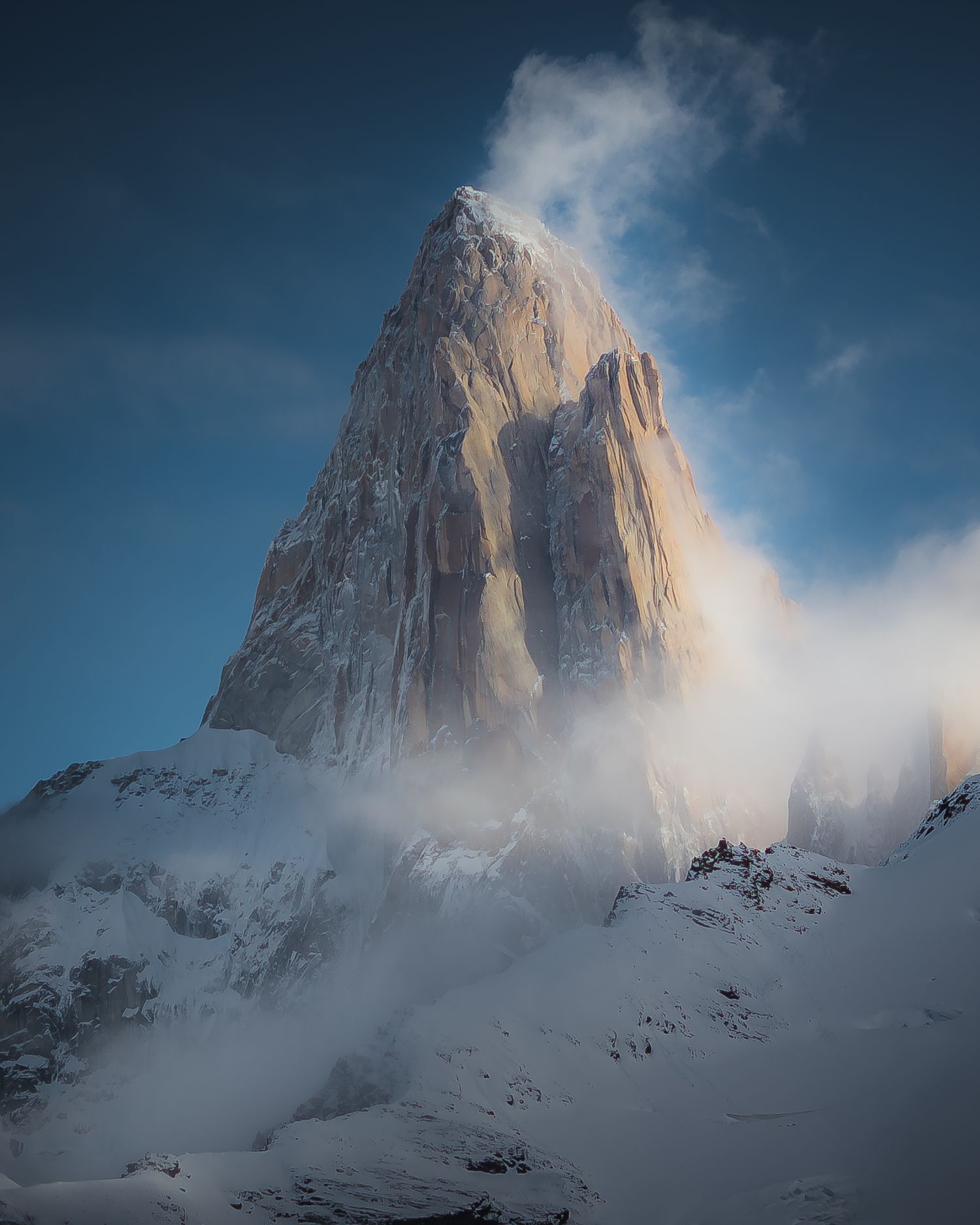 Snow-covered mountain peak with dramatic clouds swirling around its rocky summit against a blue sky.
