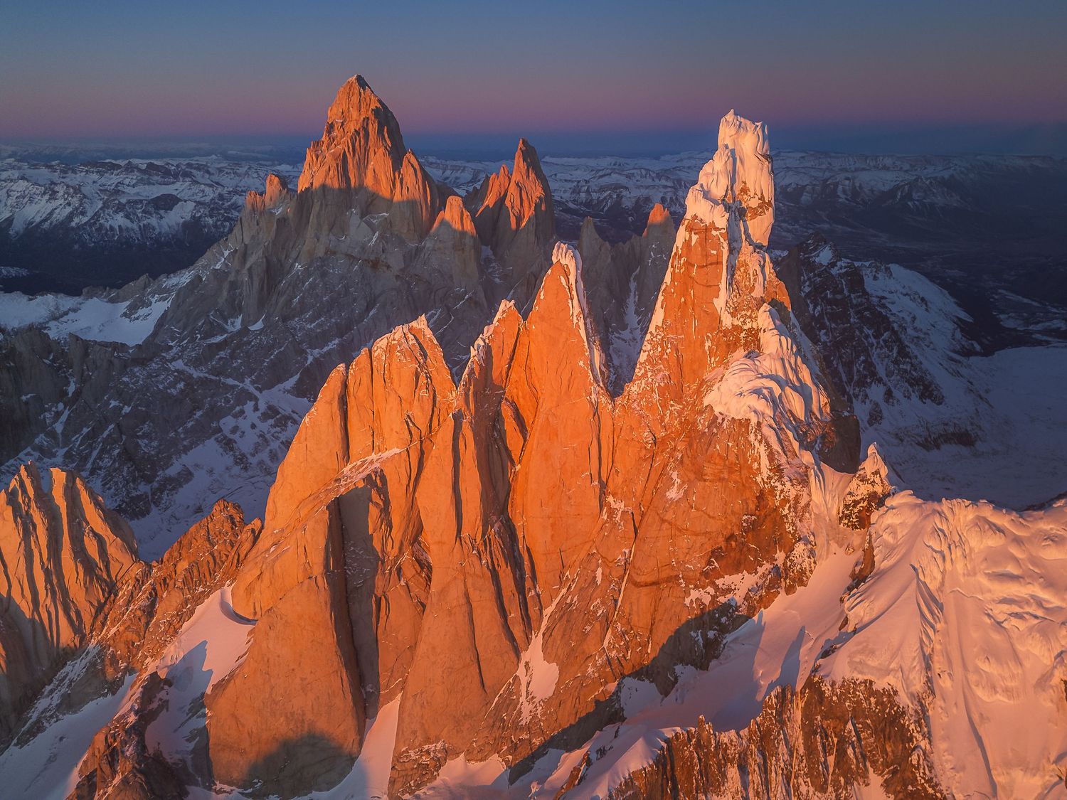 Aerial view of jagged snow-capped granite peaks glowing orange at sunset in Patagonia, Argentina.