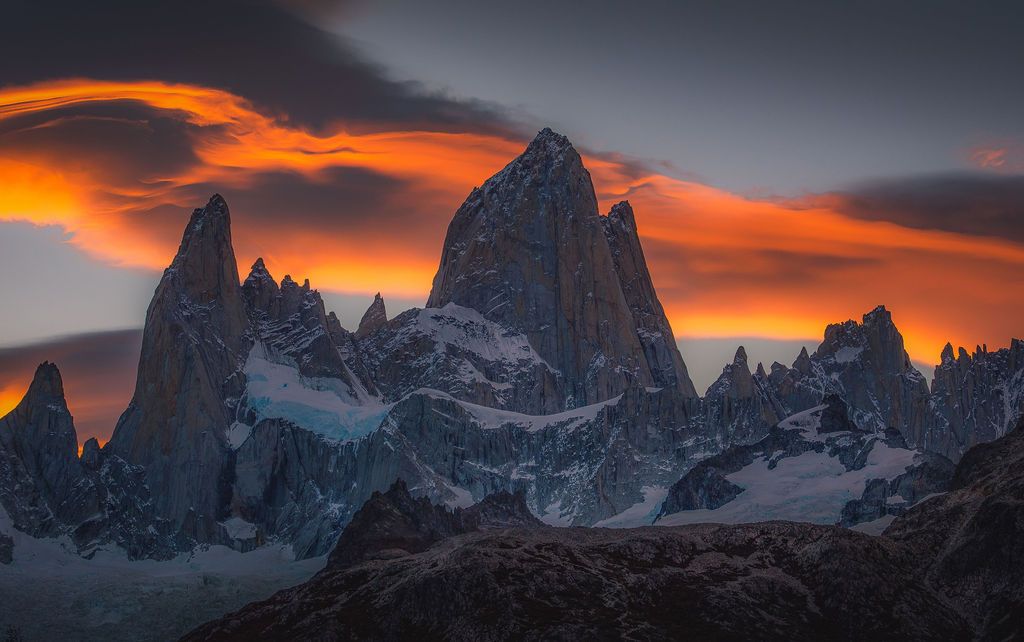 Mount Fitz Roy's sharp granite towers glow beneath dramatic orange storm clouds at sunset.