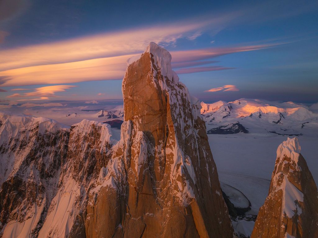 A towering ice-capped granite spire glows in warm sunset light above Patagonian glaciers and peaks.