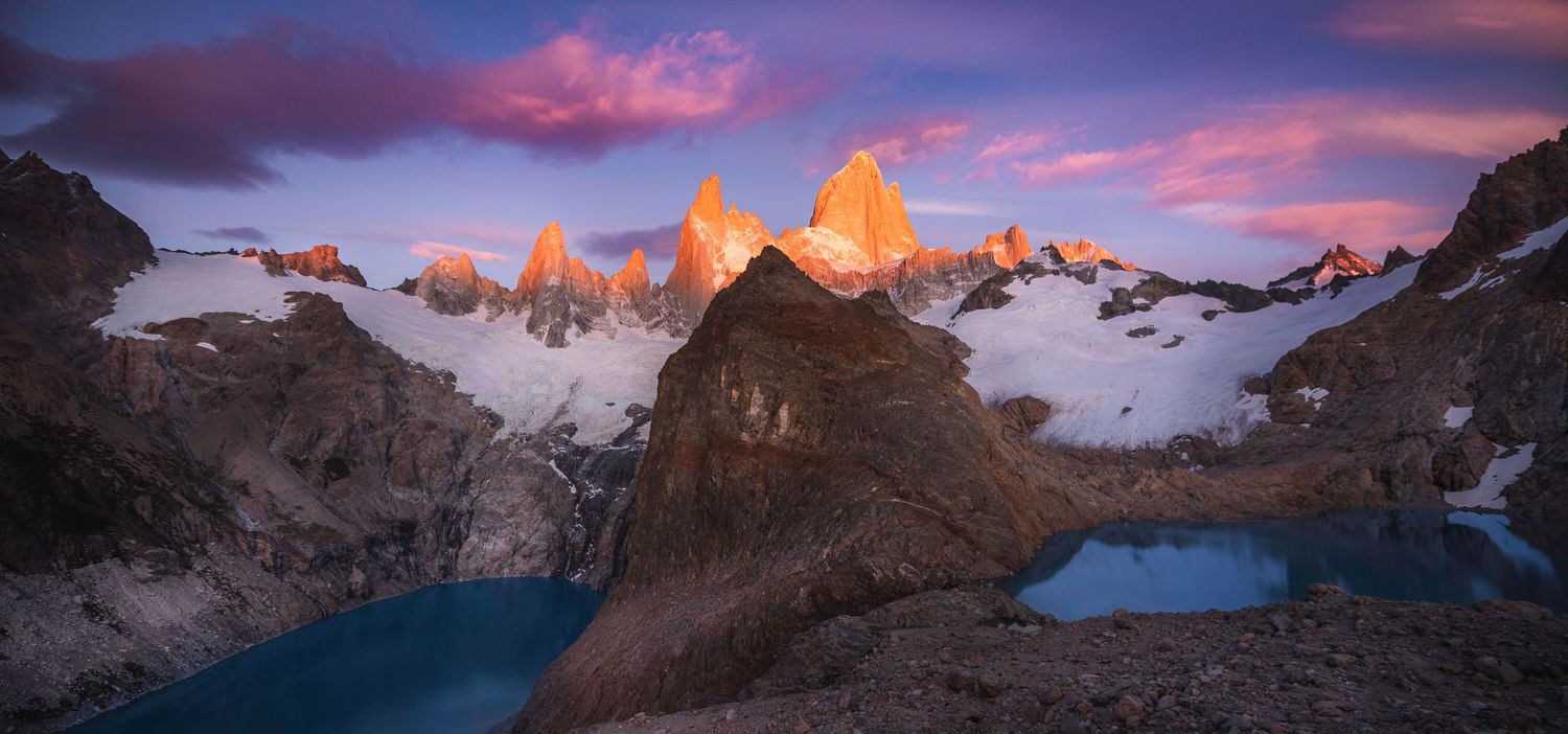 Dramatic sunrise over Mount Fitz Roy in Patagonia, with glowing pink peaks, snow, and turquoise lakes below.