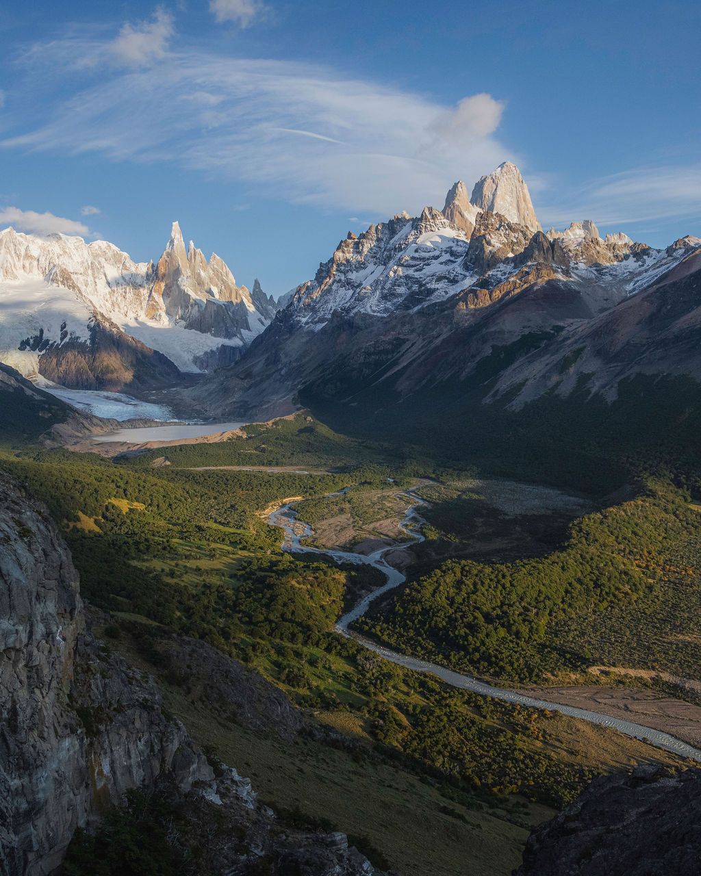 Aerial valley view of Cerro Torre and Fitz Roy massif with winding river through lush green forest.