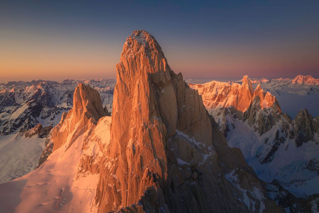 Warm alpenglow illuminates a dramatic granite spire surrounded by snow-covered peaks at golden hour.